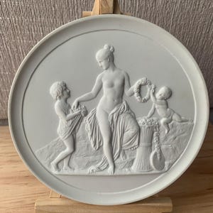 Könnte beinhalten: Eine weiße, runde Flachreliefplatte mit klassischen Figuren. Eine zentrale weibliche Figur wird von zwei kleineren Figuren flankiert, eine hält Blumen und die andere einen Kranz. Die Platte wird auf einer kleinen Holzstaffelei ausgestellt.