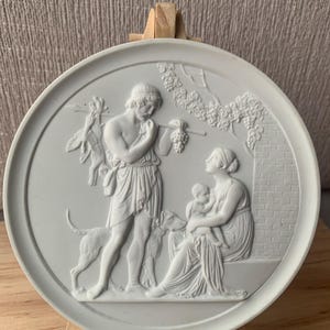 Op de afbeelding: Een ronde, lichtgrijze bas-reliëfplaat met klassieke figuren. Een man met een hond, een vrouw met een baby en druivenranken zijn afgebeeld. De plaat is gemonteerd op een kleine houten ezel.