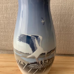 Peut inclure: Un vase en céramique bleu et blanc avec une scène d'oiseaux. Le vase présente un fond de ciel bleu clair avec un oiseau blanc en vol et un oiseau plus grand atterrissant près d'un nid. Le vase est sur une surface en bois.