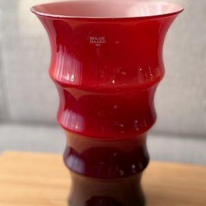 Può includere: Un vaso in vetro rosso vivace con bordo svasato e una silhouette a coste che ricorda il bambù. L'interno del bordo è di una tonalità rosa pallido. Un adesivo bianco sulla parte superiore riporta il testo HOLME GAARD 1825 in carattere sans-serif.