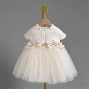 May include: A cream-colored baby dress with a tulle skirt and a matching bolero jacket. The dress features two satin bows at the waist and is adorned with small floral embellishments. The bolero has a scalloped edge and is decorated with similar floral accents.