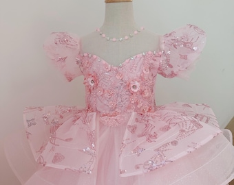 May include: A pink formal dress with a layered skirt and floral embellishments. The bodice features intricate embroidery and sparkling accents. The dress has short, puffy sleeves and is displayed on a mannequin.