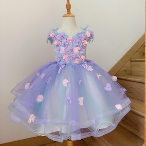 Puede incluir: Un vestido azul claro y lavanda con adornos florales. El corpiño presenta un grupo de flores rosas y moradas. La falda está en capas con tul y adornada con acentos de flores rosas dispersas. El vestido se exhibe en un maniquí.