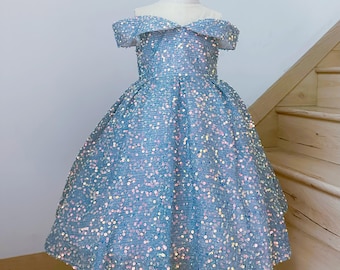 May include: A light blue off-the-shoulder dress covered in iridescent sequins. The dress has a fitted bodice and a full skirt. The dress is displayed on a mannequin. The dress is suitable for a special occasion.