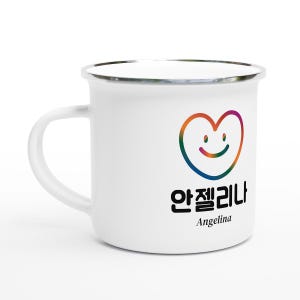 Könnte beinhalten: Weißer Emaillebecher mit silbernem Rand und großem Henkel. Der Becher zeigt ein farbenfrohes herzförmiges Design mit einem lächelnden Gesicht und dem Namen "Angelina" auf Koreanisch und Englisch.