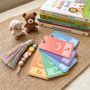 May include: A collection of children's books, including "My First Arabic Book", and flashcards with Arabic letters. A crochet teddy bear and sheep are also in the image. A tassel keychain with wooden beads and a fox charm is also present. The books are stacked.