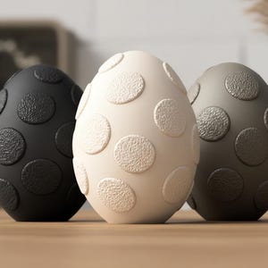 Può includere: Tre oggetti decorativi a forma di uovo. Uno è nero, uno bianco sporco e uno grigio. Ogni uovo ha una superficie strutturata con motivi circolari in rilievo. Le uova sono disposte su una superficie di legno.