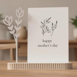 Può includere: Un biglietto bianco per la festa della mamma con illustrazioni floreali e la scritta "happy mother's day" è esposto in un supporto decorativo. Una scultura floreale in metallo bianco si trova a sinistra. Il supporto e la scultura sono su una superficie di legno.