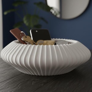 Può includere: Una ciotola bianca a coste contenente vari oggetti. Contiene uno smartphone, chiavi, monete e un piccolo oggetto in pelle. La ciotola è su una superficie di legno scuro, con una parete blu e uno specchio sullo sfondo.