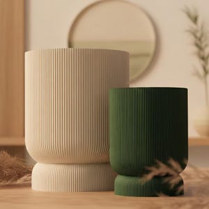 Può includere: Due fioriere cilindriche, una beige e una verde scuro, con dettagli a coste verticali. Le fioriere hanno una base arrotondata e una parte superiore abbinata. La fioriera beige è più grande di quella verde. Lo sfondo include uno specchio rotondo e una superficie in legno.