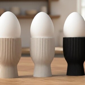 Può includere: Tre portauova nei colori neutri: beige, bianco sporco e nero. Ogni portauovo contiene un uovo bianco. I portauova hanno un design a coste e una base svasata, appoggiati su una superficie di legno. Lo sfondo è sfocato.