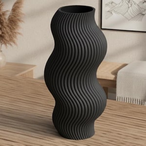 Può includere: Un vaso nero a forma di onda con scanalature verticali. Il vaso ha un'ampia apertura e una base bulbosa. È posto su un tavolo di legno. Il vaso è un oggetto decorativo, adatto per esporre fiori o come opera d'arte a sé stante.