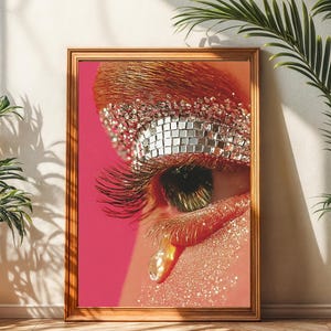 Puede incluir: Una impresión enmarcada que presenta un primer plano de un ojo glamuroso con largas pestañas y maquillaje dorado y plateado brillante. La obra de arte está sobre un fondo rosa vibrante, con un marco dorado. La imagen se muestra en una habitación con plantas.