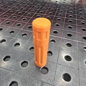 Puede incluir: Un objeto cilíndrico naranja con una parte superior texturizada y un diseño acanalado vertical. El objeto está de pie sobre una superficie metálica con agujeros espaciados uniformemente. El color es un naranja brillante y sólido.