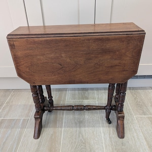 May include: A dark brown wooden drop-leaf table with a rectangular top and two hinged side leaves currently in the folded position. The table features four turned legs connected by a central horizontal stretcher and small casters at the base.