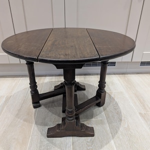 May include: A dark brown, round, drop-leaf table with a pedestal base. The table has a three-section tabletop and four turned legs. The table is approximately 76 cm in diameter when fully extended.