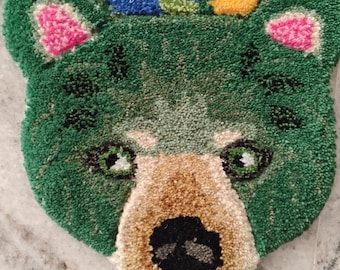 May include: A tufted rug shaped like a bear head with green fur, pink inner ears, and a tan muzzle. The bear features black eyes and a black nose. Above the bear, the rug displays a floral pattern in blue, pink, and yellow. The text Urbanwoolrugs is visible.