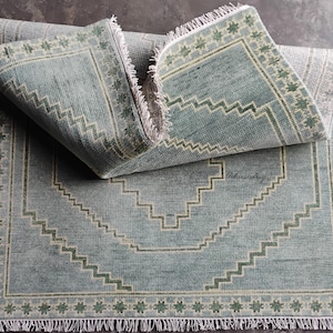 May include: A rectangular, pale blue rug with a geometric design. The rug features a repeating zigzag pattern in a lighter shade of blue and green, with a border of small floral shapes. The rug has a fringe on the edges and the text "Urbanwrig" is visible.