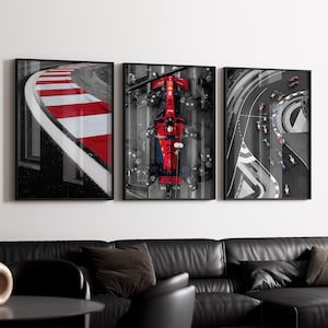 May include: Three framed prints depicting race car scenes. The left print shows a race track curve with red and white stripes. The center print features a red race car from above. The right print shows race cars on a track. The prints hang above a black leather sofa.