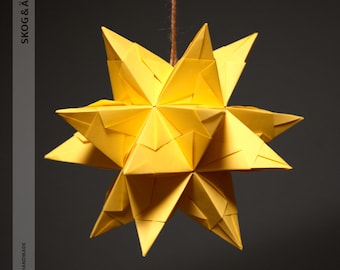 Könnte beinhalten: Hängender Stern-Dekoration aus Origami-Papier in leuchtendem Gelb. Der Stern besteht aus gefaltetem Papier und hat mehrere Spitzen. Der Text "SKOG & ÄLV" und "100% HANDMADE" sind vertikal auf der linken Seite aufgedruckt.