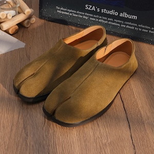 Op de afbeelding: Een paar olijfgroene schoenen met split-teen en een suède-achtige textuur. De schoenen hebben een laag profiel en een zwarte zool. De schoenen worden gepresenteerd op een houten oppervlak. De tekst "SZA's studio album" is zichtbaar.