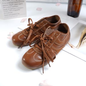 Könnte beinhalten: Ein Paar braune Ledersneaker mit weißen Nähten und Schnürsenkeln. Die Schuhe haben ein Retro-Design mit Wildlederakzenten und einer runden Spitze. Die Schnürsenkel sind gebunden.
