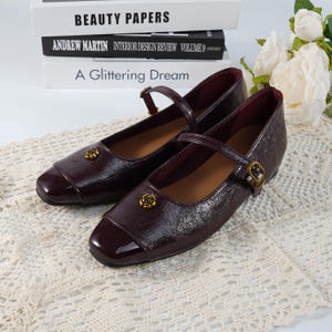 May include: Burgundy Mary Jane shoes with a glossy finish. They have a strap with a buckle and a small gold rose emblem on the toe. The shoes are displayed on a white lace surface. The shoes are a classic style.