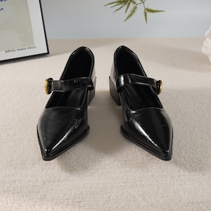 以下が含まれることがあります： ポインテッドトゥとブロックヒールが特徴の、黒いメリージェーンスタイルの靴のペア。各靴は、金色のバックルが付いたストラップが付いています。靴は光沢のある素材で作られています。ヒールは約5cmです。