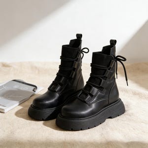 May include: A pair of black leather combat boots with chunky soles. The boots feature a lace-up design with multiple straps and a rounded toe. The boots are displayed on a neutral-coloured surface.