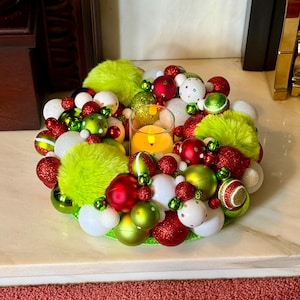May include: A circular holiday wreath centrepiece featuring a mix of red, lime green, and white ornaments in various sizes and textures, including fuzzy lime green pom-poms, with a clear glass candle holder in the centre containing a yellow LED light.