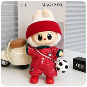 May include: A plush rabbit doll wearing a red beanie, a red football jersey with the number 7 and a crest, and matching red shorts. The doll has large eyes, a wide grin, and carries a small black and white football on a white strap.