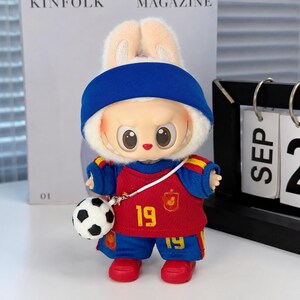 May include: A plush rabbit doll wearing a blue headband, a red and blue football jersey with the number 19 in yellow, and matching blue shorts with the number 19. The doll has a small white and black football attached by a white cord.