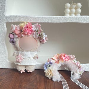 May include: A plush doll wearing a white dress and a floral crown with pink, purple, and yellow flowers. A matching floral headband with a ribbon is next to the doll. A white bubble candle sits on a shelf.