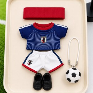 May include: A miniature football kit set comprising a blue striped shirt with a Japan crest, white shorts with red trim, a red headband, a pair of black shoes, and a small white and black football keychain with a white loop attachment.