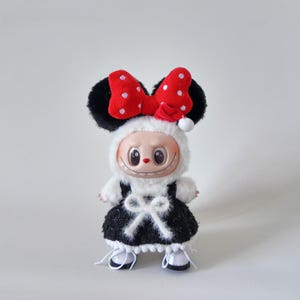 May include: A plush doll wearing a black and white dress with a white bow, white shoes, and a red bow with white polka dots. The doll has black ears and a smiling face. The doll is wearing a white furry hood.