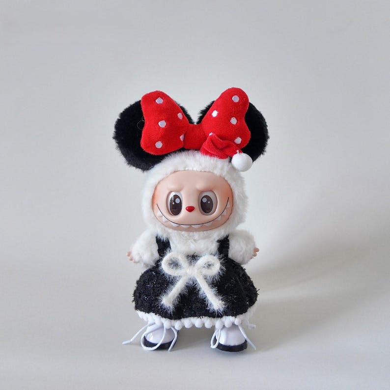 May include: A plush doll wearing a black and white dress with a white bow, white shoes, and a red bow with white polka dots. The doll has black ears and a smiling face. The doll is wearing a white furry hood.