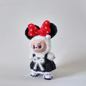 May include: A whimsical doll dressed in a black and white outfit with a large red bow with white polka dots. The doll has a smiling face and is wearing a fluffy white hood and black ears.