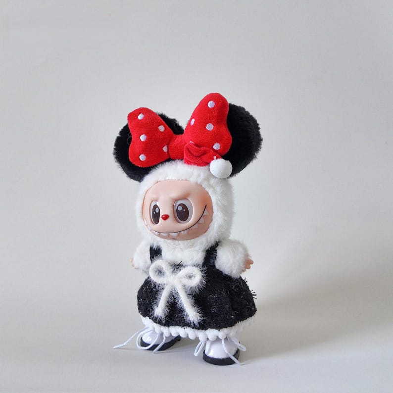 May include: A whimsical doll dressed in a black and white outfit with a large red bow with white polka dots. The doll has a smiling face and is wearing a fluffy white hood and black ears.