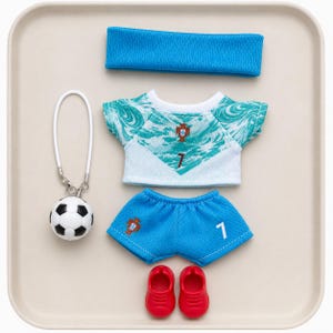 May include: A doll-sized football kit comprising a blue headband, a white and teal patterned jersey featuring the number 7, blue shorts with the number 7 and a crest, a pair of red shoes, and a small football keyring with a white loop.