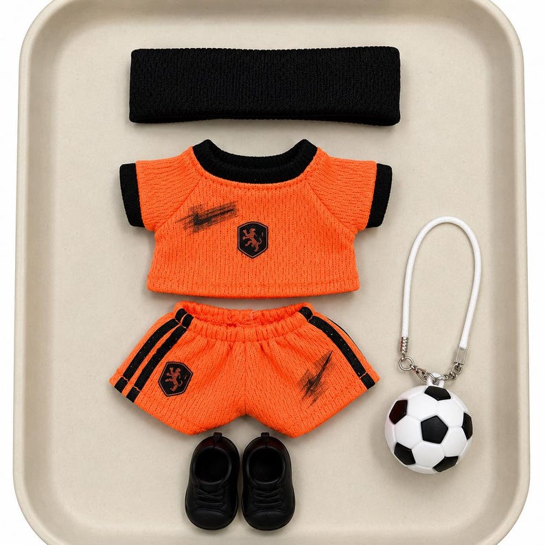 May include: A doll-sized sports outfit featuring an orange mesh t-shirt with black trim and a lion emblem, matching orange shorts with black side stripes and a lion emblem, a black headband, black shoes, and a small football keychain.