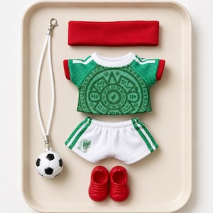 May include: A doll-sized football kit comprising a green jersey featuring a circular Aztec-style graphic, white shorts with green side stripes and a small emblem, a red headband, a pair of red shoes, and a football attached to a white lanyard.