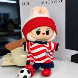 May include: A plush rabbit doll with large eyes and a wide grin, wearing a red headband, a red and white striped shirt with a small shield emblem, and dark blue shorts. The doll has red shoes and is positioned next to a small football.