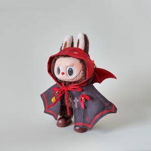 May include: A plush toy with a unique design, featuring a red hooded cape with bat-like wings and a matching red hood. The toy has a brown body, large eyes, and brown bunny ears. The cape is dark gray with red trim and decorative crosses.