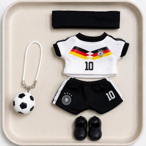 May include: A white and black football kit set comprising a jersey featuring the number 10 and German flag colours, black shorts with the number 10, a black headband, a pair of black shoes, and a football keychain with a white loop attachment.