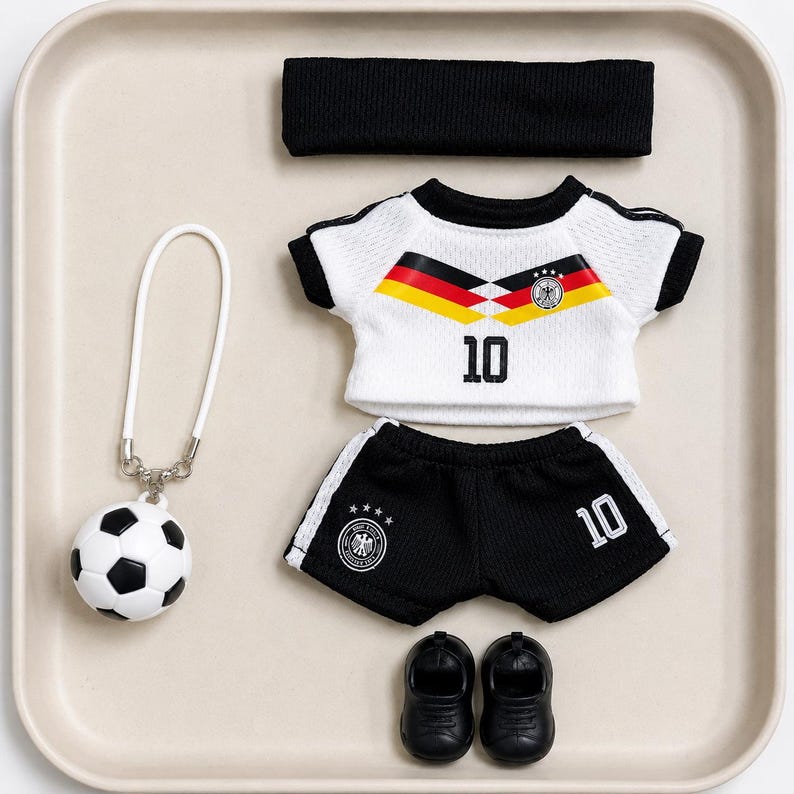 May include: A white and black football kit set comprising a jersey featuring the number 10 and German flag colours, black shorts with the number 10, a black headband, a pair of black shoes, and a football keychain with a white loop attachment.