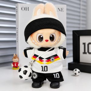 May include: A plush rabbit doll wearing a black beanie and a white football jersey with the German flag colours, black shorts with the number 10, and a small football accessory attached by a white strap. The doll has large eyes and sharp teeth.