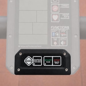 May include: A light gray Assault Fitness console with a digital display area, featuring labels for Rest, Work, Programs, and Functions. A black control panel at the bottom includes buttons labeled Enter, Comp Mode, Start, Pause, Stop, and Hold to Reset.