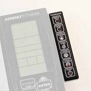 May include: A gray AssaultFitness console featuring a central LCD screen and a vertical black control panel on the right. The panel includes buttons for Interval 20-10, 10-20, Custom, Target Time, Distance, Calories, and HR. Bottom buttons include Start, Stop, and Enter.