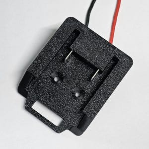 May include: A black, textured plastic battery adapter mount featuring a rectangular base with two circular mounting holes, a central slot with two metal contact terminals, and one red and one black wire extending from the top edge.