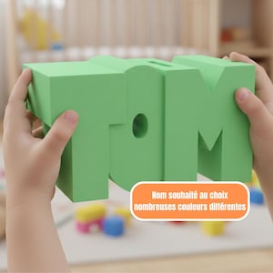 May include: A bright green foam block spelling out the name "TOM". The letters are thick and blocky, with a hole in the letter "O". The block is held by two hands. Text on the bottom right says "Nom souhaité au choix nombreuses couleurs différentes".